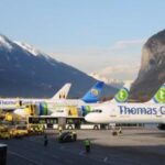 Drei Flugzeuge von Thomas Cook sind auf einem Flughafen geparkt, mit Bodenpersonal und Fahrzeugen in der Nähe, vor einer Kulisse von Bergen unter einem klaren Himmel.