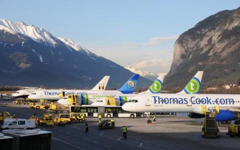 Drei Flugzeuge von Thomas Cook sind auf einem Flughafen geparkt, mit Bodenpersonal und Fahrzeugen in der Nähe, vor einer Kulisse von Bergen unter einem klaren Himmel.