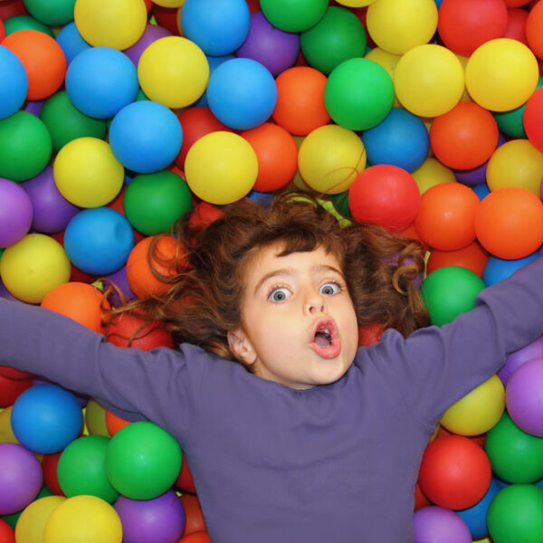 Ein Kind mit großen Augen liegt mit ausgestreckten Armen auf einem Bett aus bunten Plastikbällen und trägt ein langärmeliges lila Hemd.
