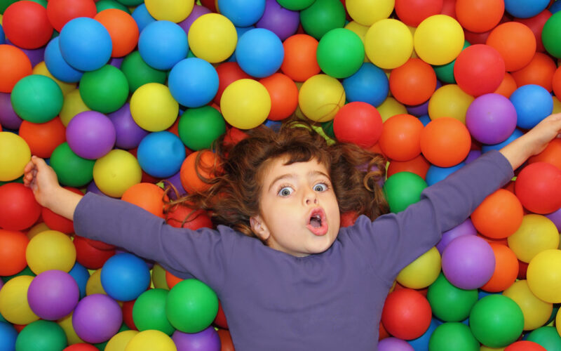 Ein Kind mit großen Augen liegt mit ausgestreckten Armen auf einem Bett aus bunten Plastikbällen und trägt ein langärmeliges lila Hemd.