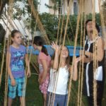 Vier Kinder und ein Erwachsener interagieren mit hängenden Seilen im Freien, mit einem Gebäude und Grünflächen im Hintergrund.