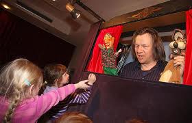 Ein Puppenspieler spielt mit zwei Puppen auf einer kleinen Bühne, während die Kinder zusehen und nach den Puppen greifen.