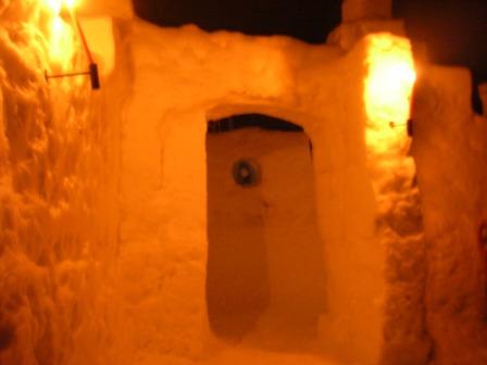 Ein Eingang aus festgefahrenem Schnee, der von warmen gelben Lichtern beleuchtet wird, führt zu einem weiteren schneebedeckten Bereich.