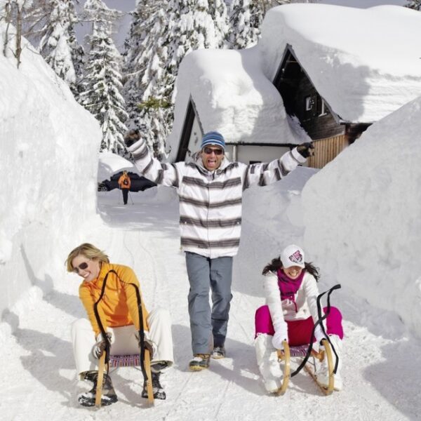 Zwei Kinder auf Schlitten sitzen auf einem verschneiten Weg, während ein Erwachsener mit erhobenen Armen hinter ihnen steht; im Hintergrund sind Schneebänke und Häuser zu sehen.