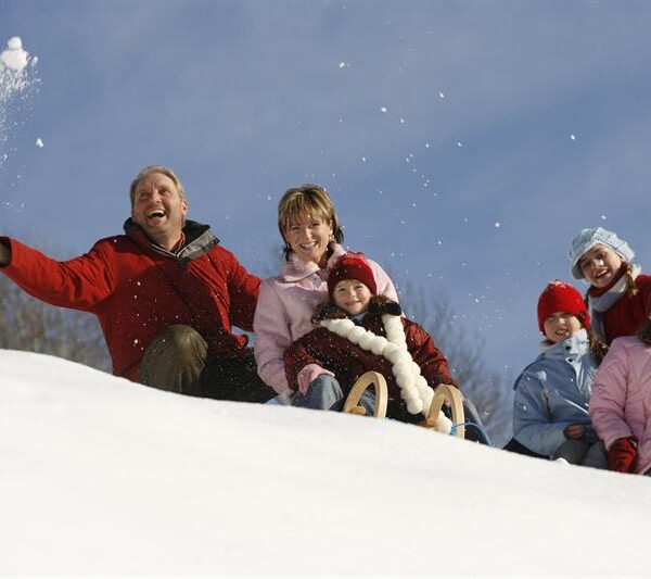 Sechs Personen, darunter Erwachsene und Kinder, sitzen in Winterkleidung auf einem verschneiten Hügel. Ein Erwachsener wirft Schnee, während ein Kind auf einem Schlitten sitzt. Der Himmel ist klar und blau.