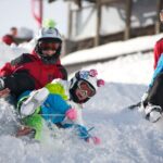 Drei Personen in bunten Winterklamotten und mit Helmen schlittern einen verschneiten Hügel hinunter und werfen dabei Schnee auf.