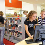 Mehrere junge Leute benutzen Computer und stöbern in den Bücherregalen einer modernen, hell erleuchteten Bibliothek.