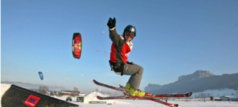 Ein Skifahrer mit Helm und Skibrille springt auf einer verschneiten Piste und hält dabei einen Griff, der an einem Drachen im Hintergrund befestigt ist.