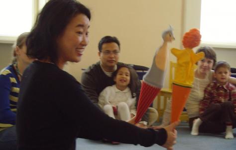 Eine Frau hält zwei Handpuppen vor einer Gruppe von sitzenden Erwachsenen und Kindern in einem Klassenzimmer hoch.