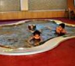 Drei Kinder schwimmen in einem kleinen, unregelmäßig geformten Hallenbad, das von einem roten Bodenbelag umgeben ist.