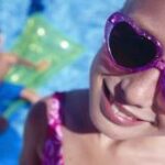 Nahaufnahme eines Kindes mit herzförmiger Sonnenbrille, das in die Kamera lächelt, mit einem anderen Kind auf einem grünen Schwimmer in einem Schwimmbad im Hintergrund.