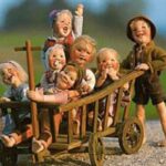 Sieben lächelnde Puppen in altmodischer Kleidung, sechs davon in einem Holzkarren sitzend und eine daneben stehend, vor einem unscharfen Hintergrund im Freien.
