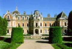 Ein großes, verziertes Herrenhaus mit vielen Fenstern und Schornsteinen, umgeben von gepflegten Hecken und einem Kiesweg unter einem strahlend blauen Himmel.
