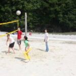 Vier Personen spielen Beachvolleyball auf einem Sandplatz, im Hintergrund sind Bäume zu sehen.
