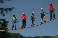 Fünf Personen mit Helmen und Klettergurten überqueren eine Hängeseilbrücke im Freien, mit Bäumen und Bergen im Hintergrund.