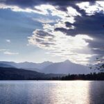 Ein ruhiger See mit Bergen im Hintergrund, unter einem teilweise wolkenverhangenen Himmel und durchbrechenden Sonnenstrahlen.