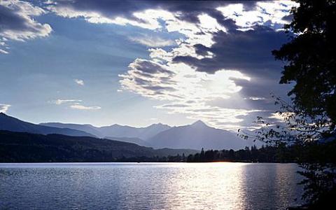 Ein ruhiger See mit Bergen im Hintergrund, unter einem teilweise wolkenverhangenen Himmel und durchbrechenden Sonnenstrahlen.