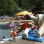 Kinder spielen in einem flachen Außenbecken mit einer roten Rutsche in Form eines Elefanten; Erwachsene und Kinder entspannen sich auf dem Rasen und den Liegestühlen in der Nähe.