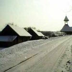 Schneebedeckte Straße, die zu einer Ansammlung von Gebäuden mit steilen Dächern und einem kleinen Glockenturm führt, umgeben von Schnee in einer ländlichen Winterlandschaft.