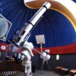 Ein großes Teleskop ist in einem kuppelförmigen Observatorium mit blauen Wänden und einer gewölbten Decke montiert. An den Wänden sind ein Bild des Mondes und Wegweiser zu sehen.