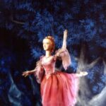 Eine Marionettenballerina in einem rosafarbenen Kostüm steht auf einem Bein vor einem blauen Blätterhintergrund mit nebelartigen Effekten zu ihren Füßen.