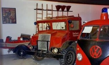 Zwei rote Oldtimer-Feuerwehrautos, eines davon mit Leiter und Löscheimern an der Wand dahinter, sind im Innenraum neben einem kleinen roten Boot und einem Volkswagen-Einsatzfahrzeug ausgestellt.