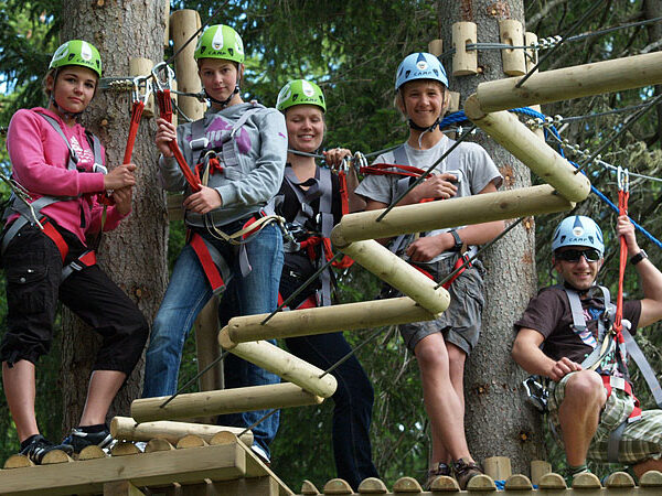 Fünf Personen mit Helmen und Gurten stehen auf einer hölzernen Plattform in einem Baumkronen-Parcours, umgeben von Bäumen und Seilhindernissen.