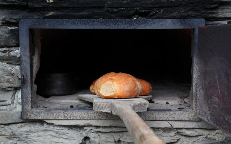 Ein Laib Brot auf einem Holzbrett wird in einen Steinofen mit einer Metalltür gelegt oder aus diesem herausgenommen.