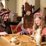 Drei Personen in mittelalterlichen Kostümen spielen ein historisches Brettspiel an einem Holztisch, mit Töpferwaren und einer Feuerstelle im Hintergrund.