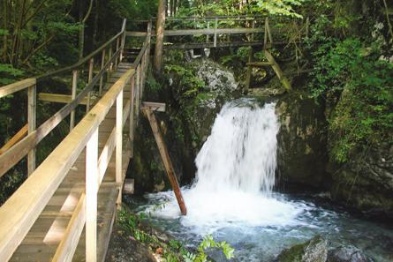 Ein kleiner Wasserfall mündet in ein felsiges, von Grün umgebenes Becken, über das ein Holzsteg und ein Fußweg führen.