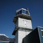 Ein weißer Flugsicherungsturm mit großen Fenstern erhebt sich vor einem strahlend blauen Himmel, im Hintergrund ist ein schneebedeckter Berg zu sehen.