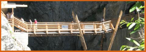 Eine hölzerne Fußgängerbrücke mit Geländer überspannt eine felsige Schlucht, auf der mehrere Personen entlanggehen.