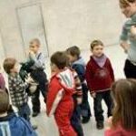 Eine Gruppe kleiner Kinder steht in einem Raum mit einer erwachsenen Frau, einige stehen mit dem Gesicht zur Wand, andere unterhalten sich oder warten.