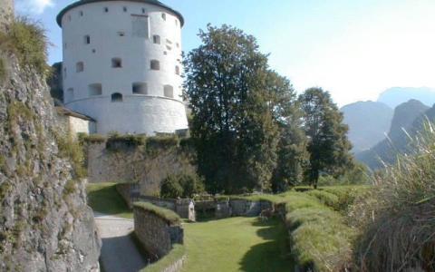 Ein runder weißer Steinturm steht auf einem grasbewachsenen Hügel, der von Steinmauern und Bäumen umgeben ist, während im Hintergrund die Berge zu sehen sind.