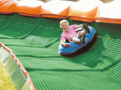 Eine Person fährt mit einem blauen aufblasbaren Schlauch eine grüne künstliche Rutsche hinunter, umgeben von orangefarbenen Polstern.