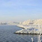 Ein ruhiger Fluss schlängelt sich durch eine frostige Landschaft mit schneebedeckten Bäumen und Felsen unter einem strahlend blauen Himmel.