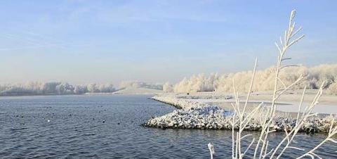 Ein ruhiger Fluss schlängelt sich durch eine frostige Landschaft mit schneebedeckten Bäumen und Felsen unter einem strahlend blauen Himmel.