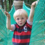 Ein Junge mit blondem Haar steht auf einer Seilbrücke im Freien und hält sich mit beiden Händen an dem grünen Netz fest.