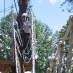 Zwei Personen mit Helmen stehen auf einer hölzernen Plattform zwischen Bäumen und bereiten sich auf das Überqueren einer Seilbrücke in einem Hochseilgarten vor.