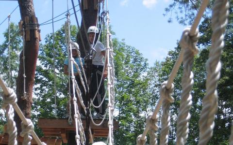 Zwei Personen mit Helmen stehen auf einer hölzernen Plattform zwischen Bäumen und bereiten sich auf das Überqueren einer Seilbrücke in einem Hochseilgarten vor.