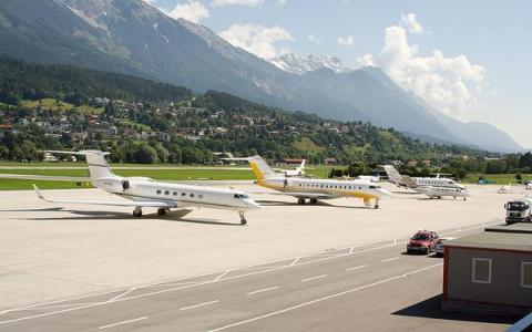 Mehrere Privatjets sind auf dem Rollfeld eines Flughafens geparkt, im Hintergrund sind Berge und eine Stadt zu sehen.