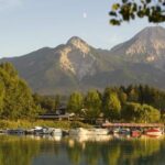 Kleine Boote, die an einem ruhigen See angedockt sind, mit Häusern und dichten Bäumen im Vordergrund und zwei hohen, baumbestandenen Bergen im Hintergrund unter einem klaren Himmel.