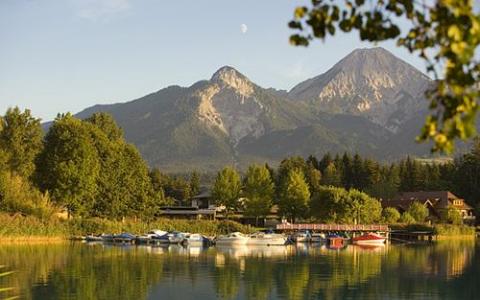 Kleine Boote, die an einem ruhigen See angedockt sind, mit Häusern und dichten Bäumen im Vordergrund und zwei hohen, baumbestandenen Bergen im Hintergrund unter einem klaren Himmel.