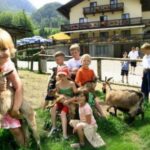 Mehrere Kinder spielen mit Ziegen auf einer Wiese in der Nähe eines Holzzauns, mit einem großen Gebäude und Bergen im Hintergrund.