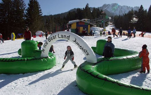 Kinder spielen auf einem verschneiten Hügel unter einem aufblasbaren Bogen mit der Aufschrift "Nici Bear Land Jungholz", während im Hintergrund Erwachsene und ein Skilift zu sehen sind.