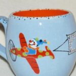Hellblaue Keramiktasse mit Henkel, die einen Cartoon-Clown zeigt, der ein rot-gelb gepunktetes Flugzeug fliegt und eine Fahne zieht.