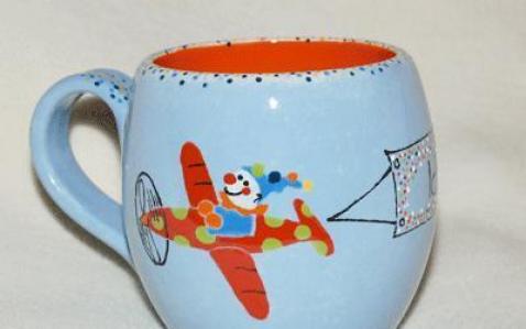 Hellblaue Keramiktasse mit Henkel, die einen Cartoon-Clown zeigt, der ein rot-gelb gepunktetes Flugzeug fliegt und eine Fahne zieht.