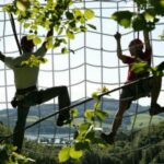 Zwei Personen mit Klettergurten klettern an einem Seilnetz im Freien, umgeben von Bäumen und einer malerischen Landschaft im Hintergrund.