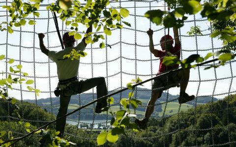Zwei Personen mit Klettergurten klettern an einem Seilnetz im Freien, umgeben von Bäumen und einer malerischen Landschaft im Hintergrund.