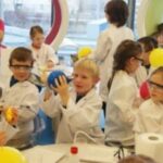 Kinder in Laborkitteln und mit Schutzbrillen nehmen an einer wissenschaftlichen Aktivität an einem Tisch im Klassenzimmer teil und hantieren mit Luftballons und verschiedenen Laborgeräten.